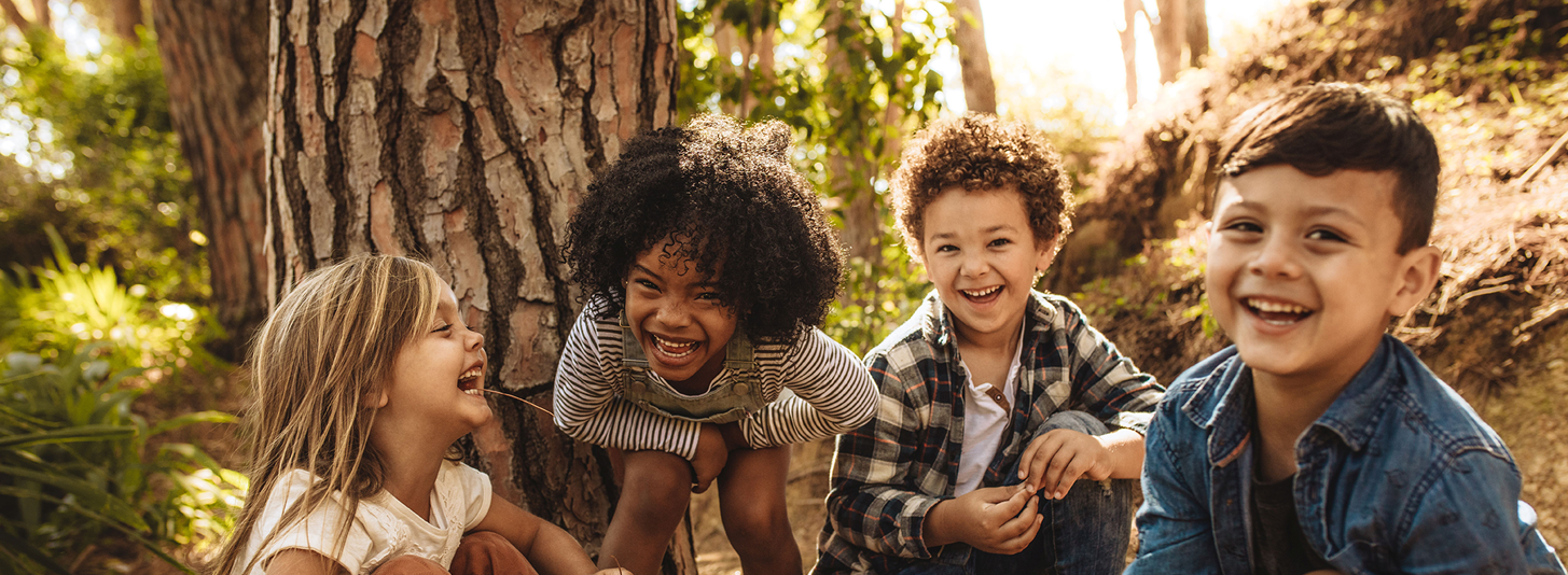 Vier Kinder im Kindergartenalter mit unterschiedlicher Hautfarben hocken im Wald und lachen in die Kamera