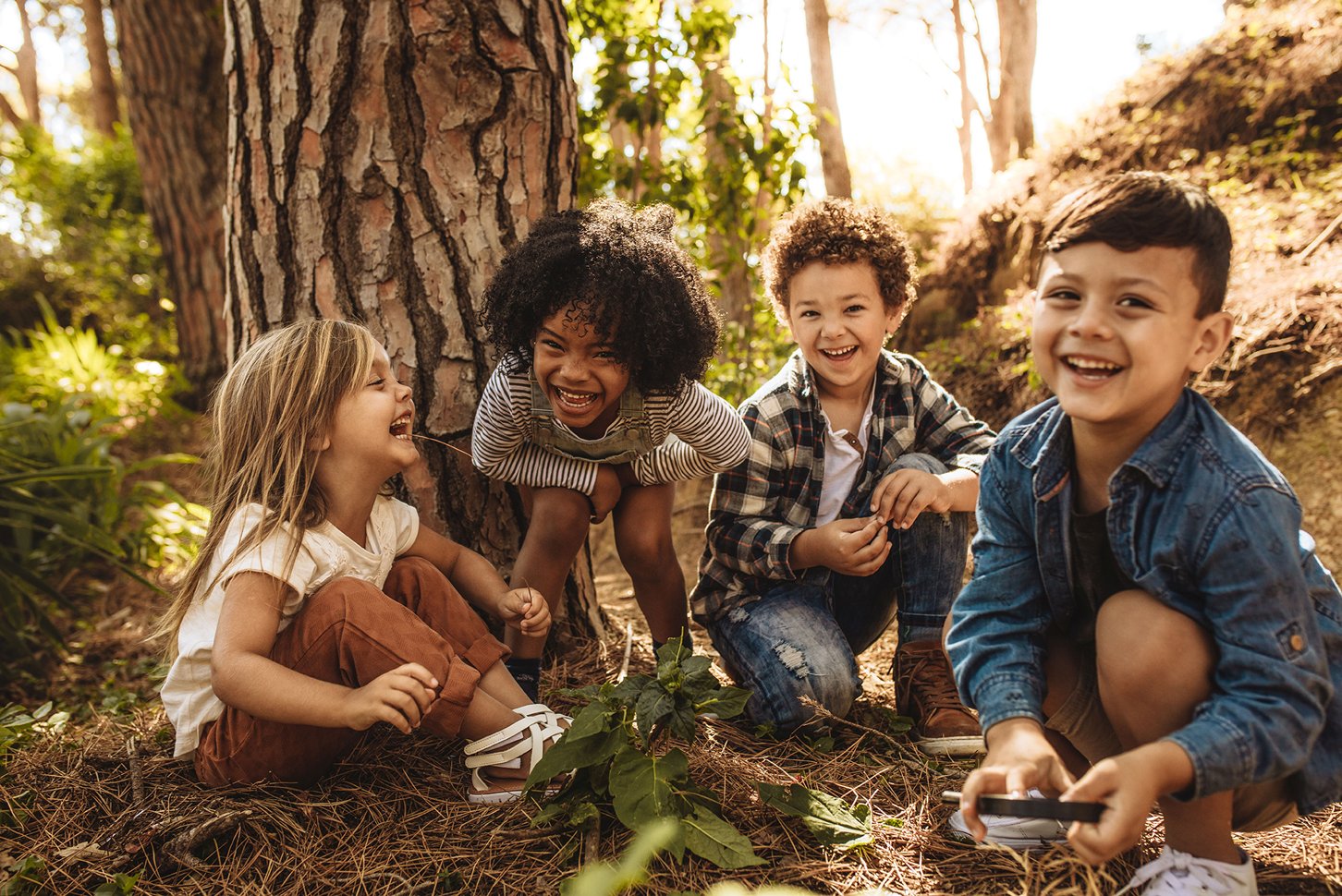 Vier Kinder im Kindergartenalter mit unterschiedlicher Hautfarben hocken im Wald und lachen in die Kamera