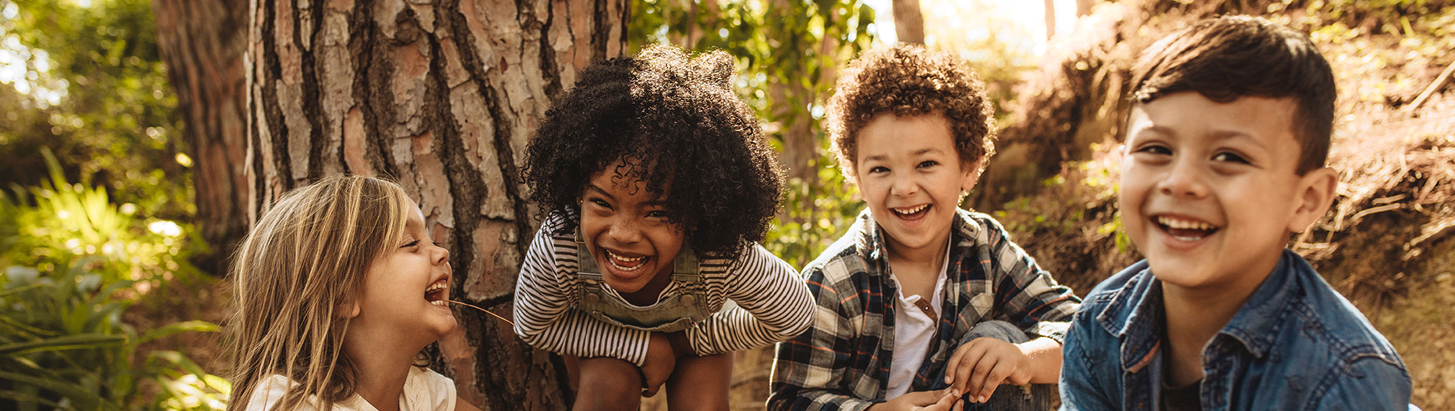 Vier Kinder im Kindergartenalter mit unterschiedlicher Hautfarben hocken im Wald und lachen in die Kamera