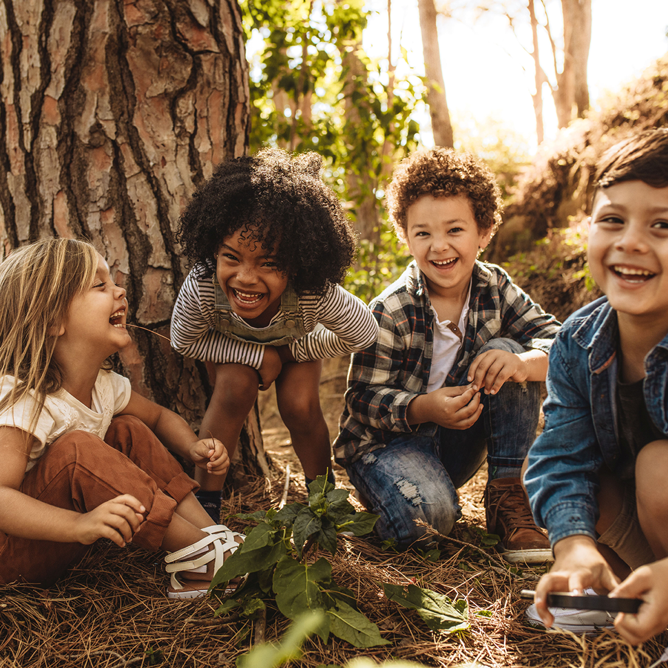 Vier Kinder im Kindergartenalter mit unterschiedlicher Hautfarben hocken im Wald und lachen in die Kamera