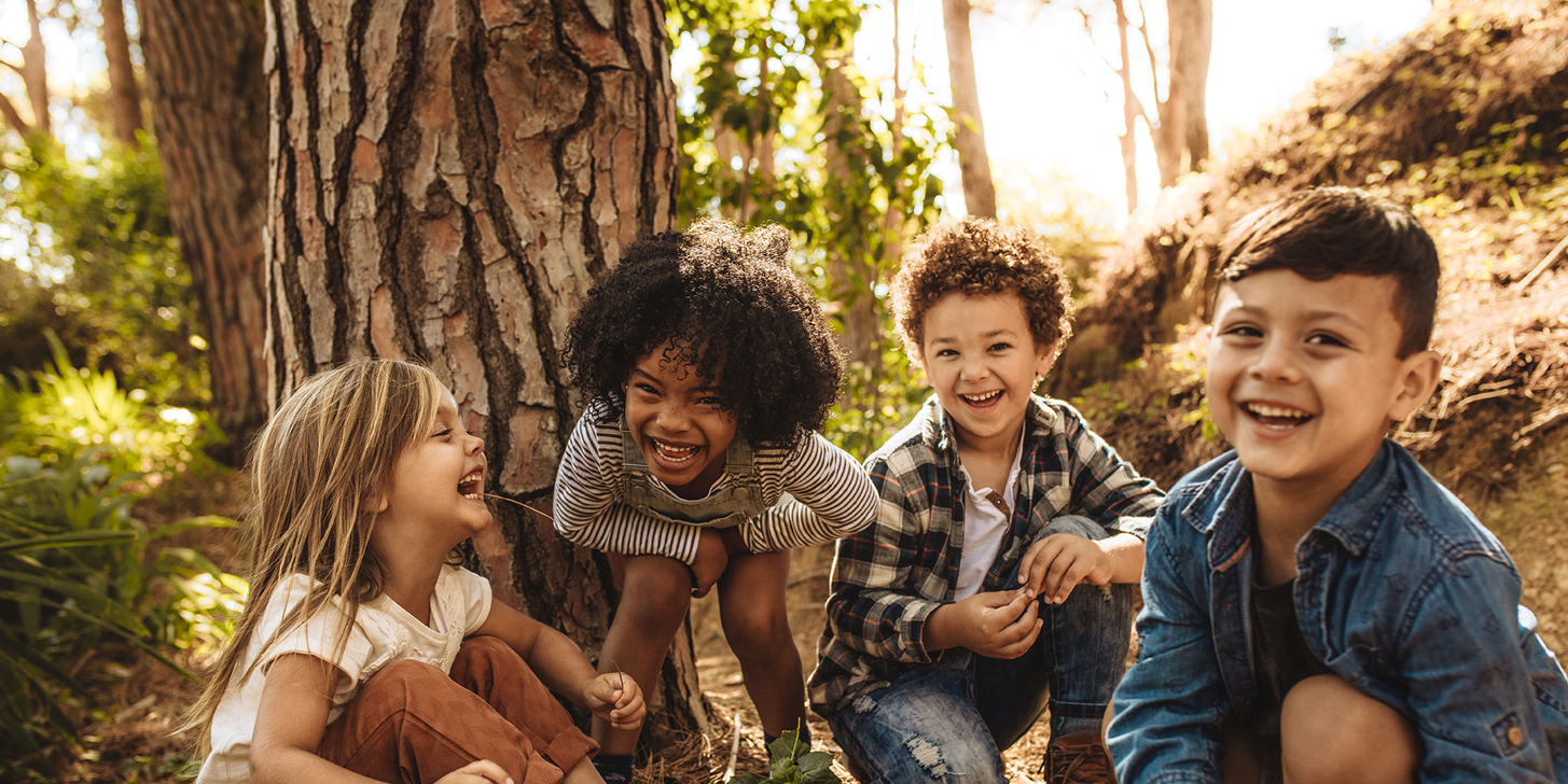 Vier Kinder im Kindergartenalter mit unterschiedlicher Hautfarben hocken im Wald und lachen in die Kamera