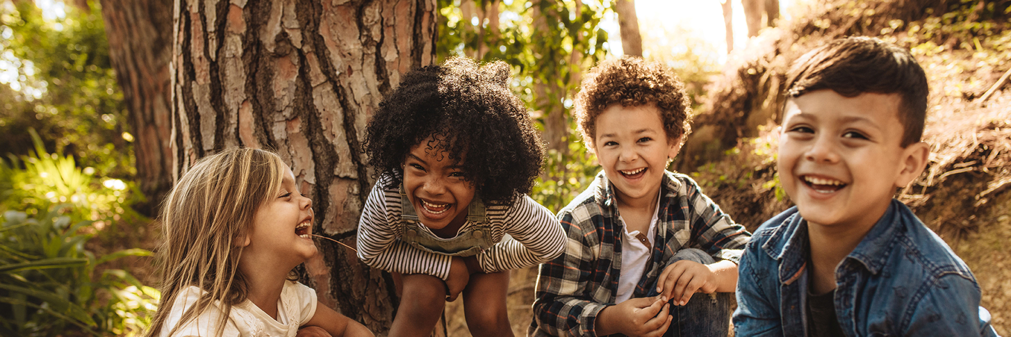 Vier Kinder im Kindergartenalter mit unterschiedlicher Hautfarben hocken im Wald und lachen in die Kamera