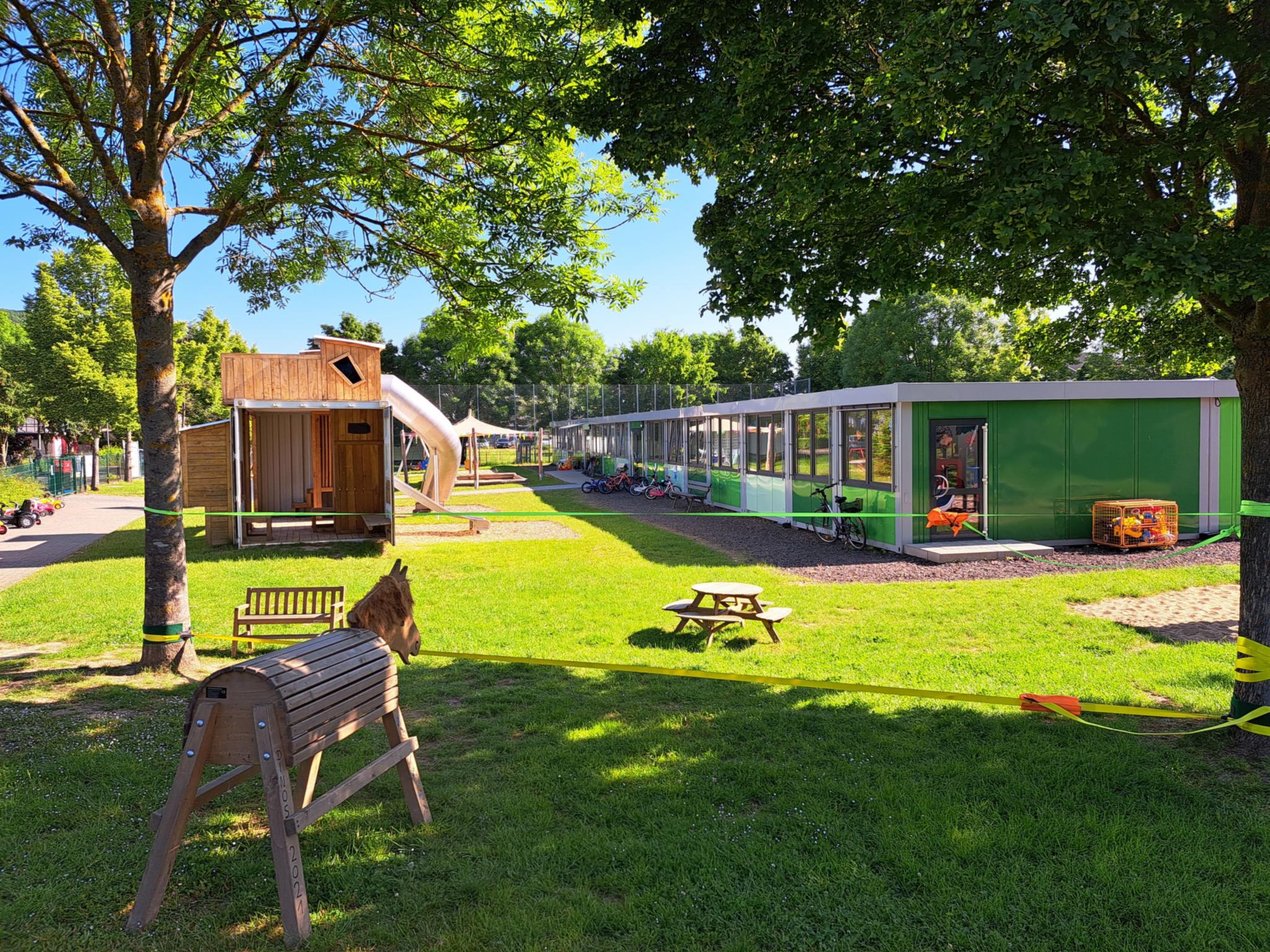 Außenanlage der Kita St. Maurititus BN-AW-Heimersheim Eine Wiese mit großen Spielgeräten, einem Holzpferd vor einem grünen, flachen, langen KiTa-Gebäude